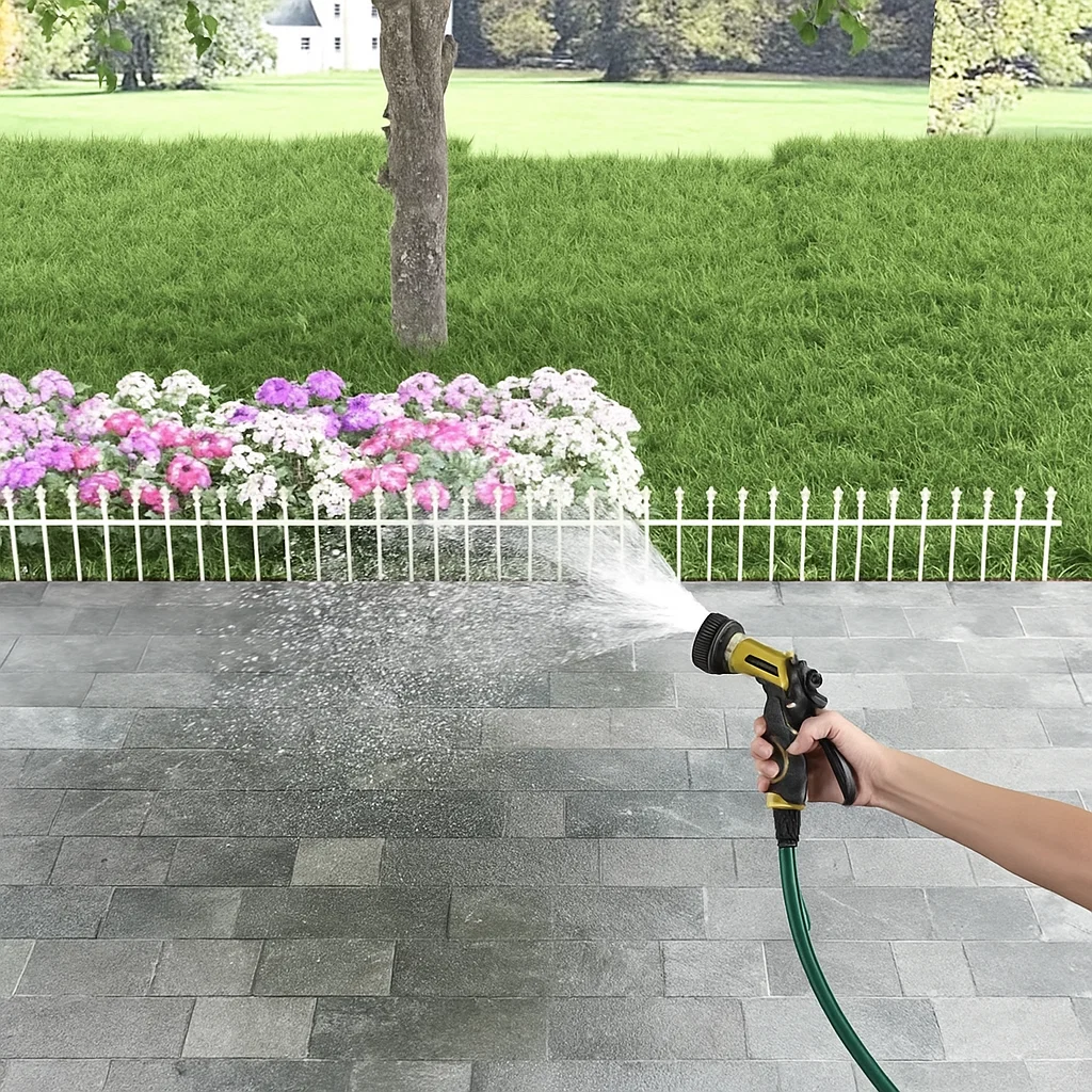 Hand holding hose spraying water on patio near white picket fence and blooming flower garden.
