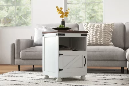 White farmhouse-style end table with open shelf and barn door, placed near grey sectional in bright room.