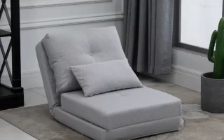 Compact light grey futon chair with pillow, placed on floral rug near grey curtain and cactus plant.