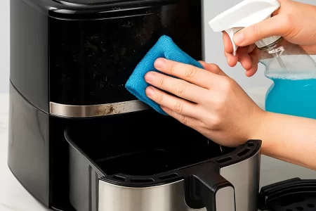 Person wiping greasy air fryer surface with cloth and spray cleaner during cleaning.