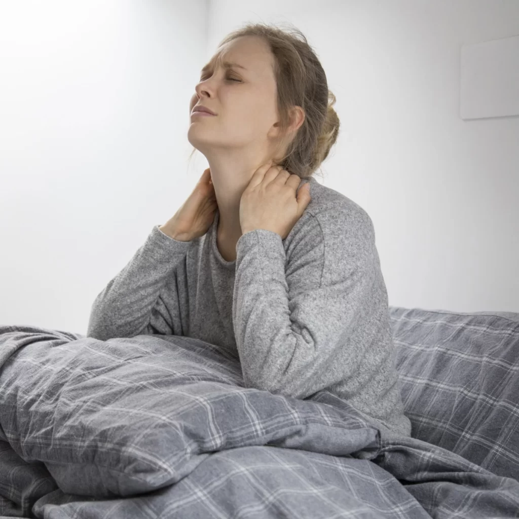 Woman sitting in bed holding neck in pain, showing discomfort from poor sleep posture.