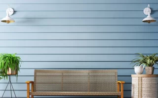 Outdoor wicker bench with wooden frame against light blue wall and indoor plants on a wooden deck.