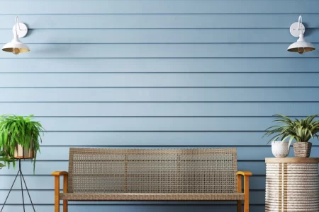 Outdoor wicker bench with wooden frame against light blue wall and indoor plants on a wooden deck.