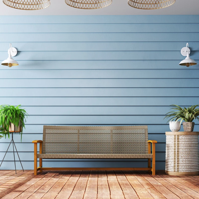 Outdoor wicker bench with wooden frame against light blue wall and indoor plants on a wooden deck.