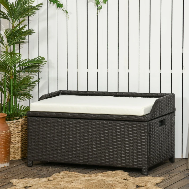 Dark brown wicker outdoor storage bench with cream cushion against white fence and tropical plants.